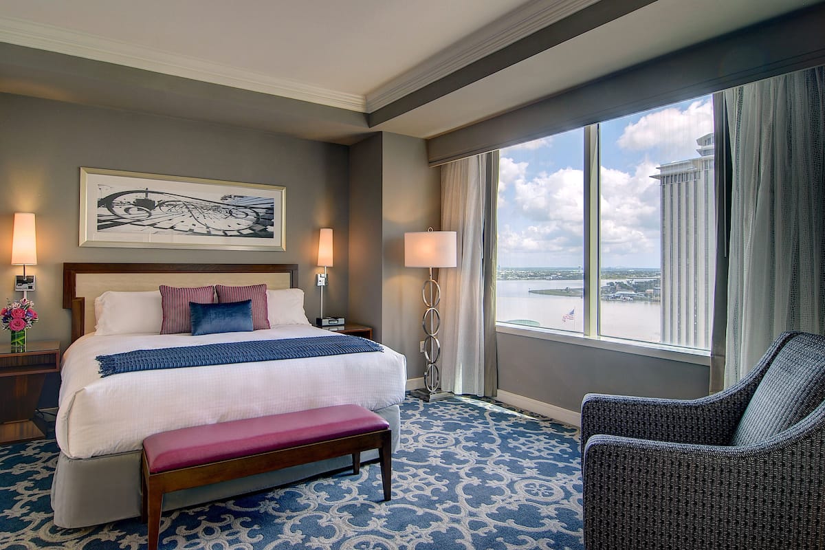 Ein Hotelzimmer mit einem großen Bett, einer Bank, einem Sessel und Blick auf die Stadt, sowie einem Gemälde an der Wand.