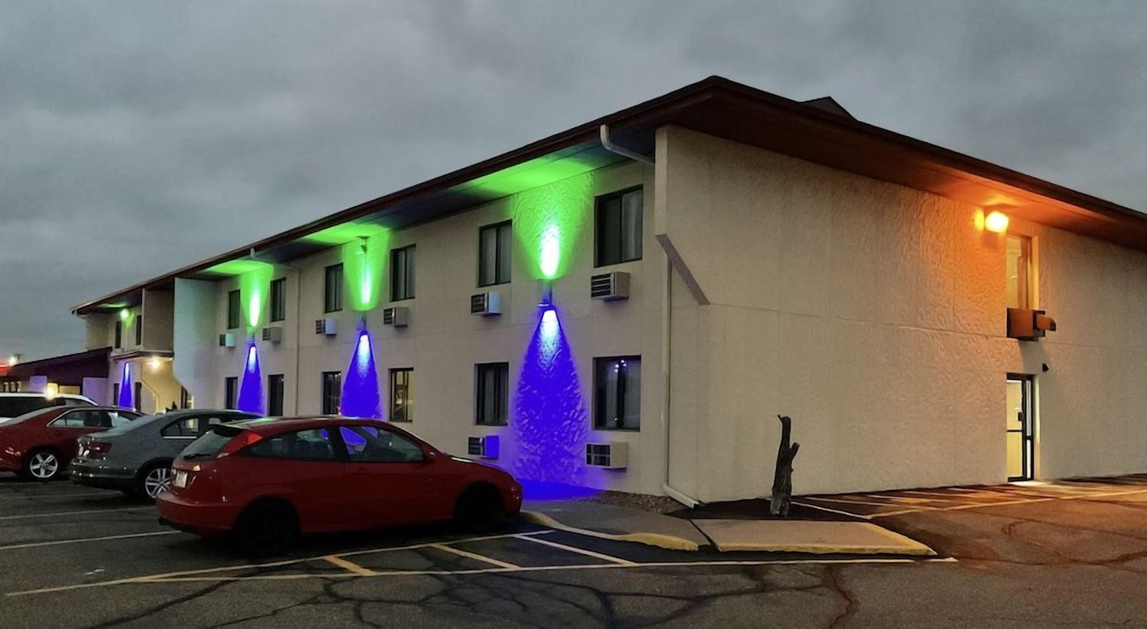 Un hôtel à plusieurs étages doté d’un parking, avec une rangée de colonnes illuminées et une voiture rouge garée au premier plan.