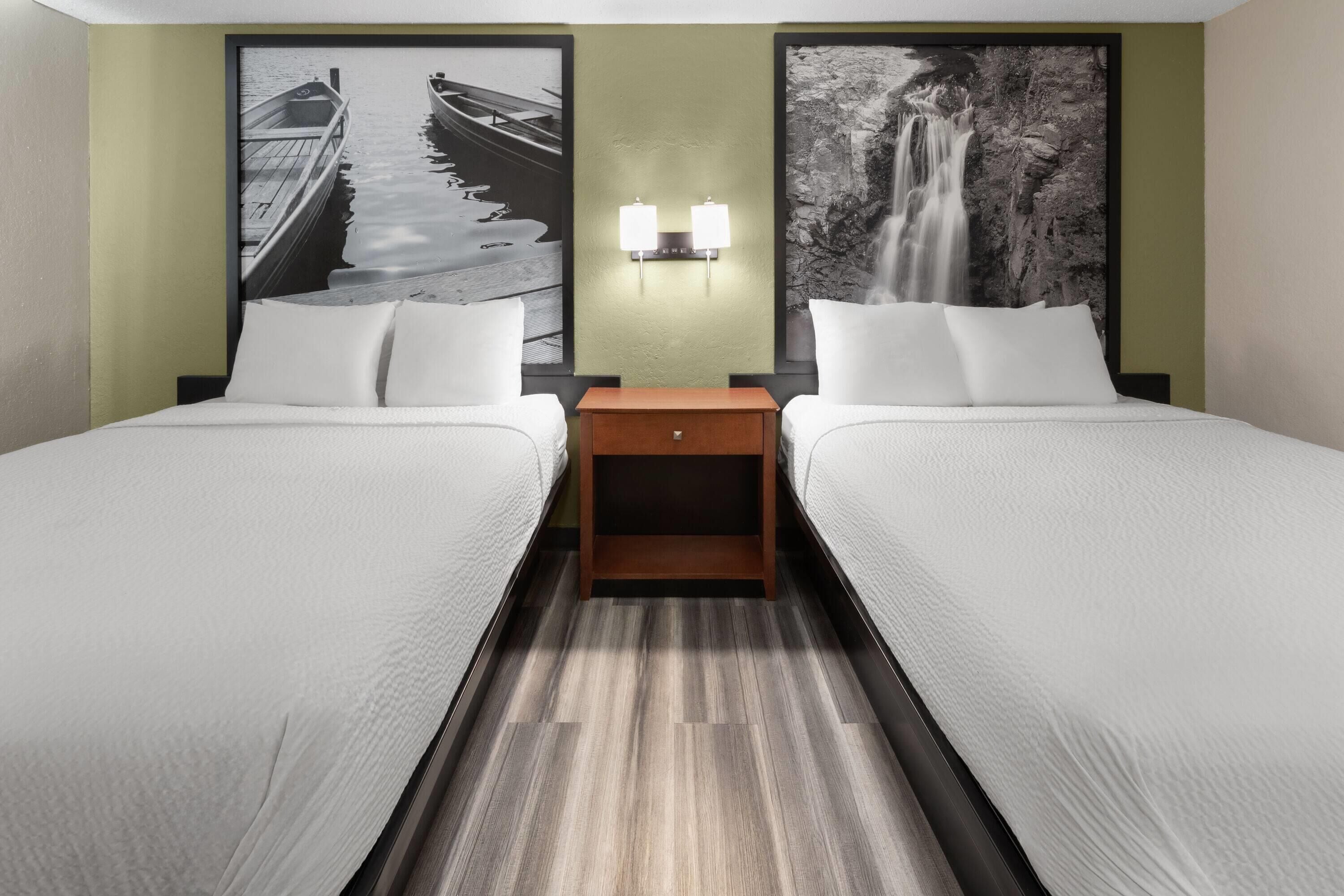Quarto de hotel com duas camas, uma mesa de cabeceira e duas luminárias fixadas na parede.