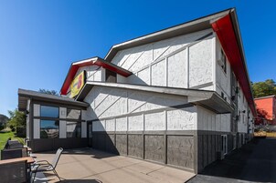Un edificio con fachada blanca y detalles en rojo, que presenta un gran ventanal y un pequeño patio con mesas al aire libre.