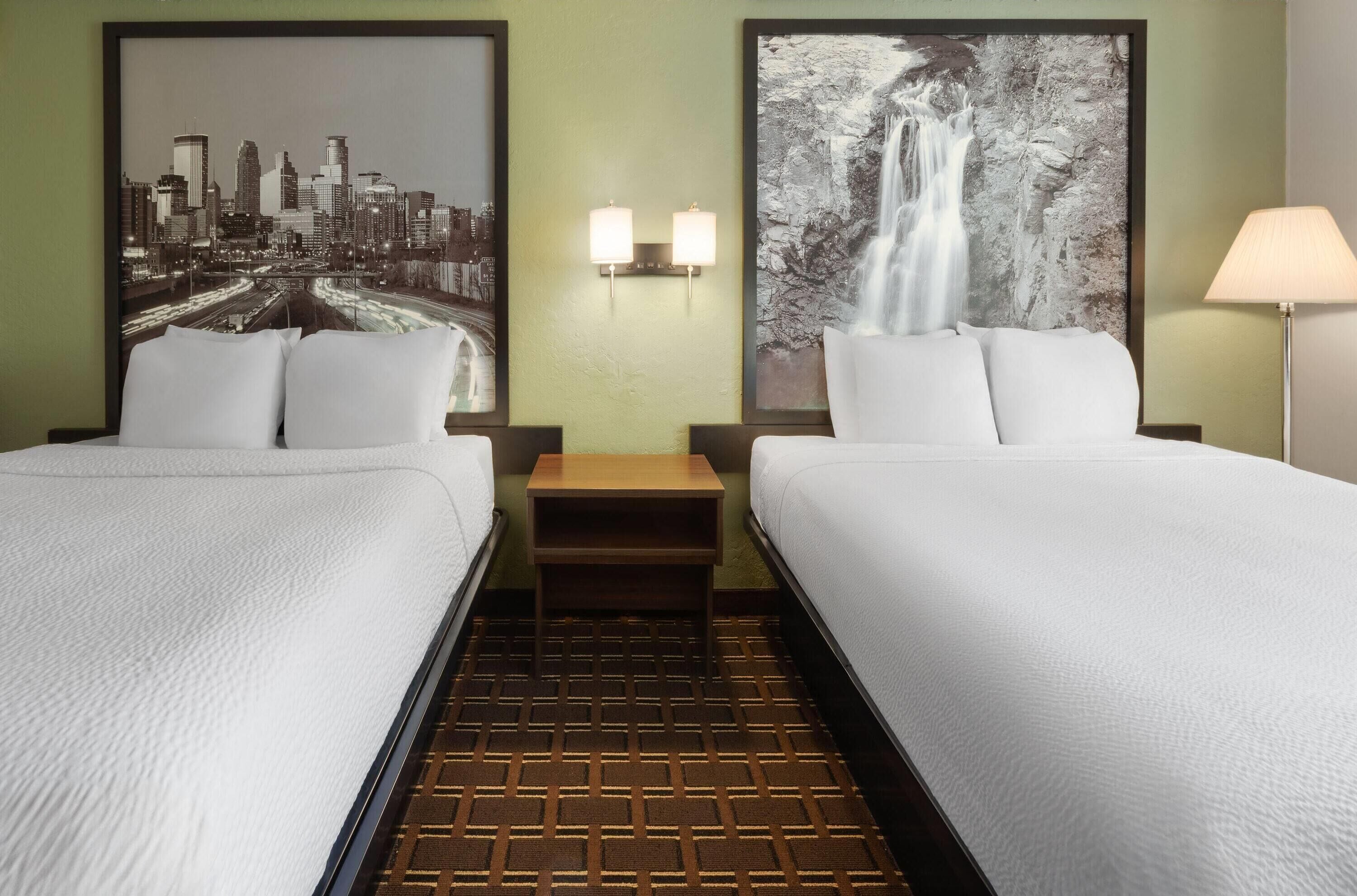 A hotel room with two beds, a cityscape and waterfall poster on the wall, a nightstand with a lamp, and a patterned carpet.