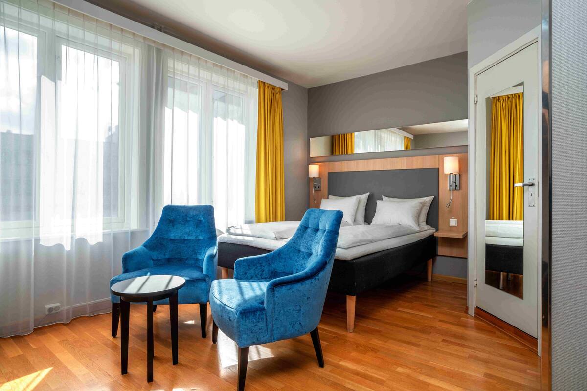 A hotel room with a bed, two blue armchairs, a small table, and a mirror above the bed.