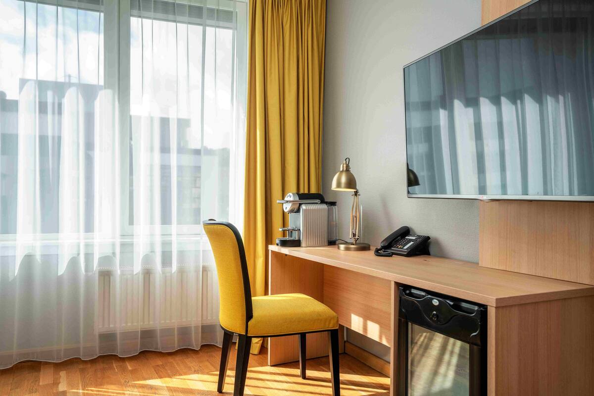 A hotel room with a wooden desk, a yellow chair, a radiator, a lamp, a phone, and a large window with sheer curtains.
