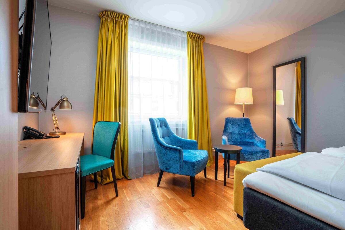 A hotel room with a bed, two chairs, a desk with a lamp, a phone, and a TV.