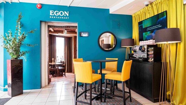 A restaurant interior with a teal wall, yellow chairs, a black table, and a TV screen. - Thon Hotel Lillestrøm (Lillestrom)