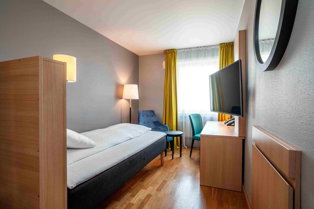 A hotel room with a bed, a desk, a chair, a TV, and a lamp.