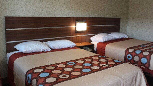 A hotel room with two beds, each with a patterned bedspread, a wooden headboard, and a bedside lamp. - American Inn (Branford)