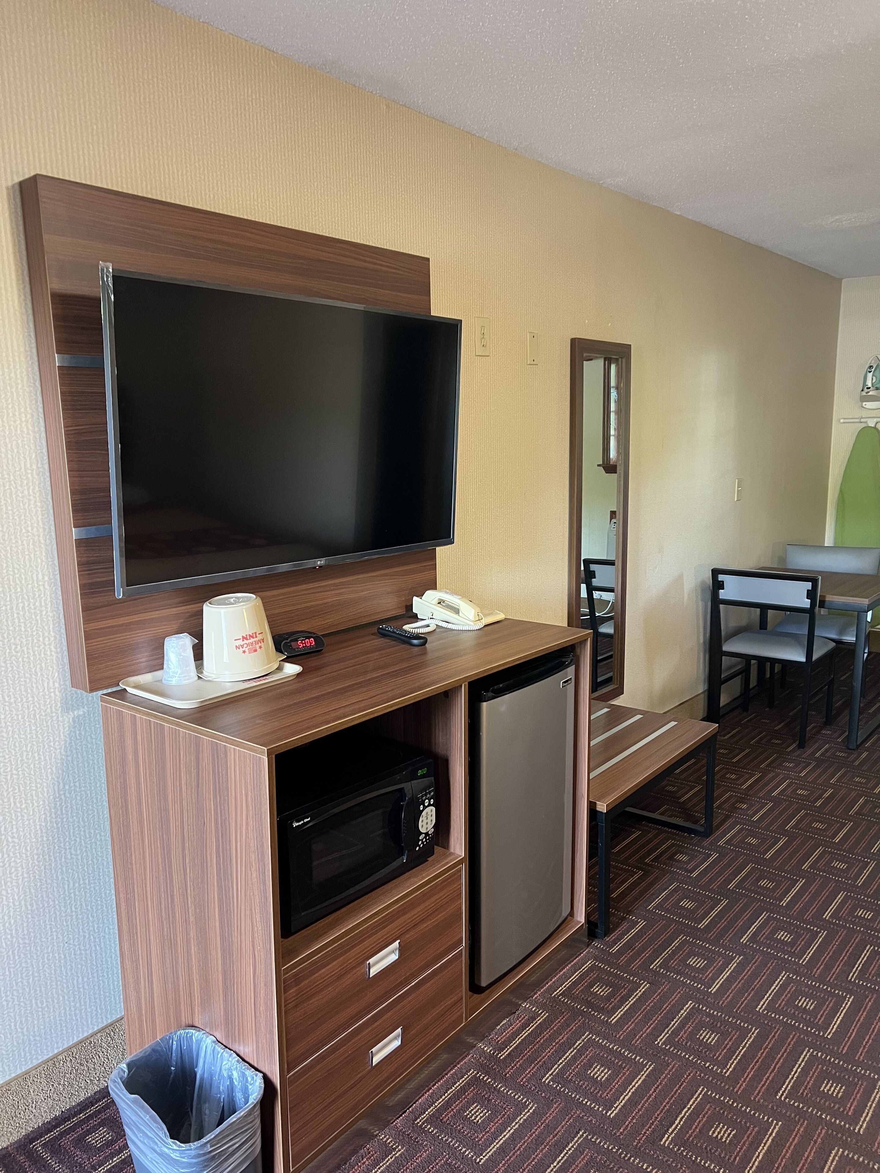 A hotel room with a flat-screen TV, a minibar, a desk with a telephone, and a mirror.