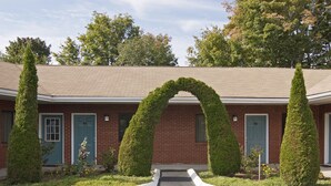 A brick building with a symmetrical archway formed by topiary trees, a front parking area, and a clear sky. - American Inn (Branford)