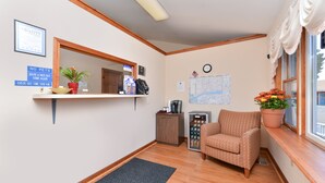A hotel reception area with a wooden floor, a reception desk, a chair, a coffee machine, a map on the wall, and a clock. - American Inn (Branford)