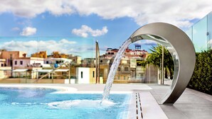 Ein moderner Pool im Freien mit Wasserspiel, umgeben von Gebäuden und blauem Himmel.