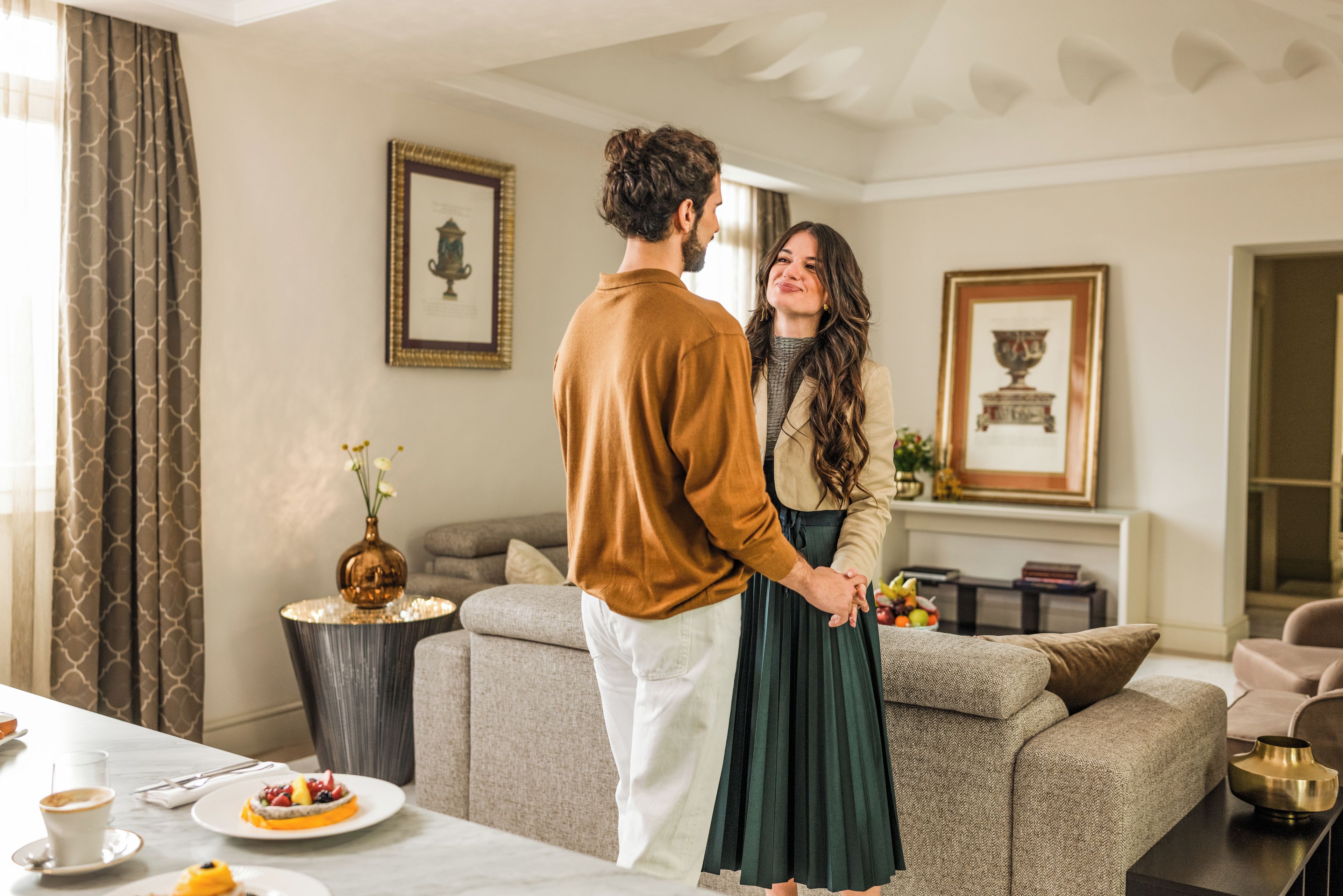 A couple standing in a living room with a sofa, a coffee table, and a dining table with a plate of fruit.