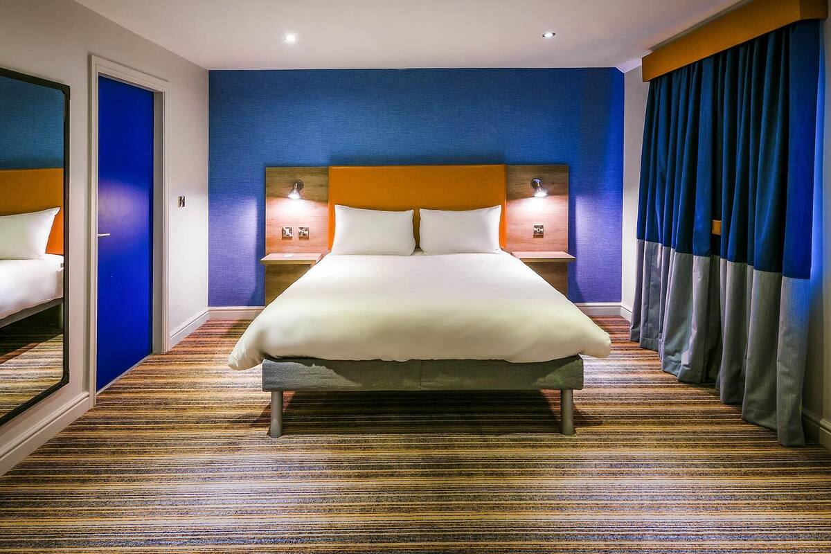 Une chambre d’hôtel avec un grand lit, deux tables de chevet avec des lampes, une porte bleue et un tapis à rayures.