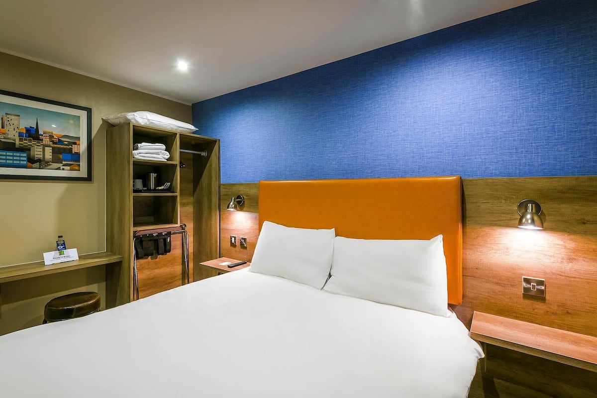 Une chambre d’hôtel avec un grand lit, une tête de lit en bois, une table de chevet, un bureau avec une chaise, une armoire et un tableau au mur.