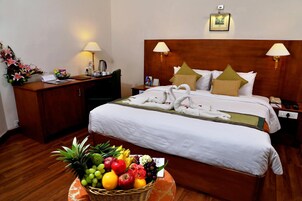 A hotel room with a neatly made bed, a bedside table with a lamp, a nightstand, a desk with a lamp, and a fruit basket on the floor.