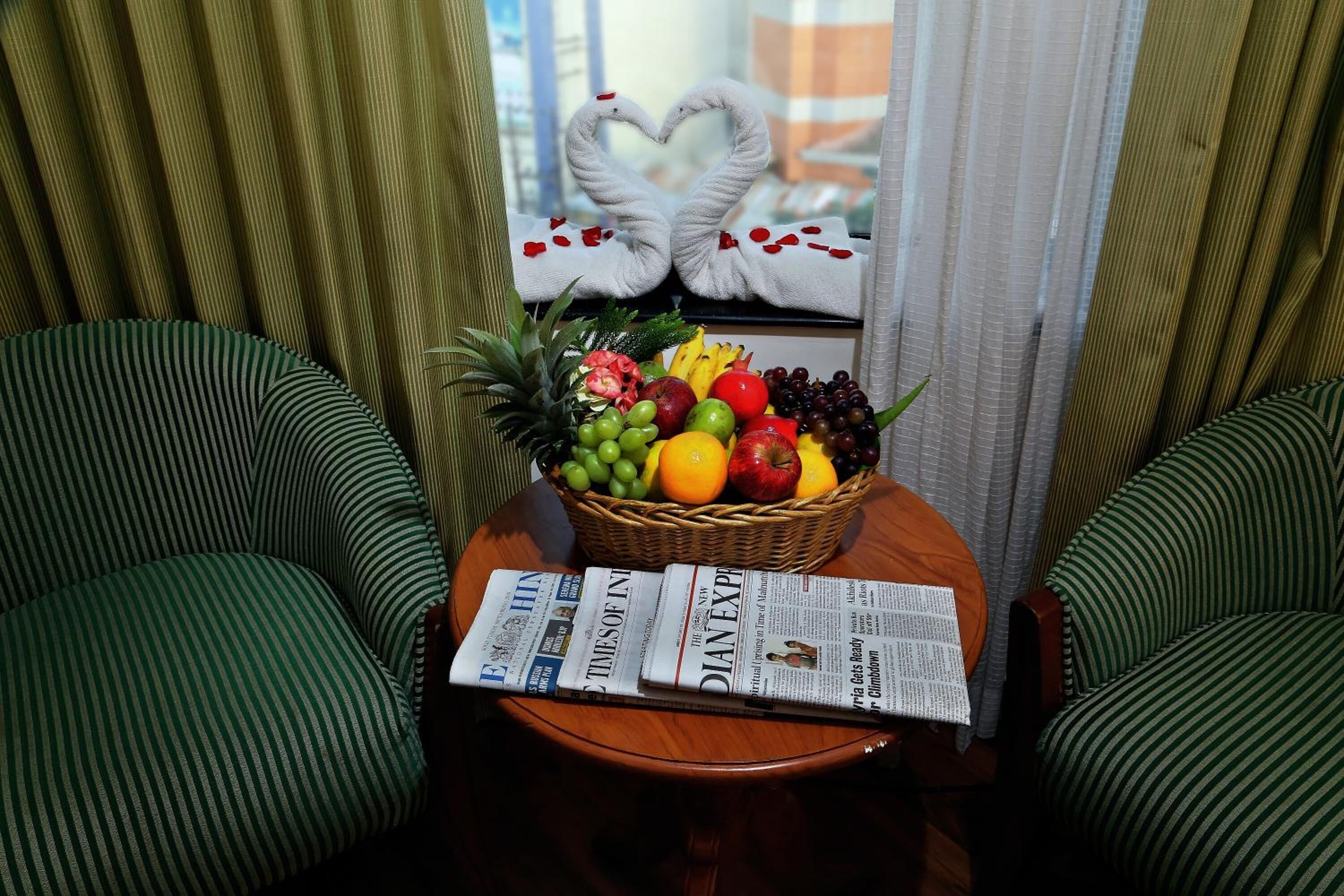 Ein Zimmer mit zwei grün gestreiften Sesseln, einem kleinen Holztisch mit Obstkorb, Zeitungen und einem Fenster mit Vorhängen.