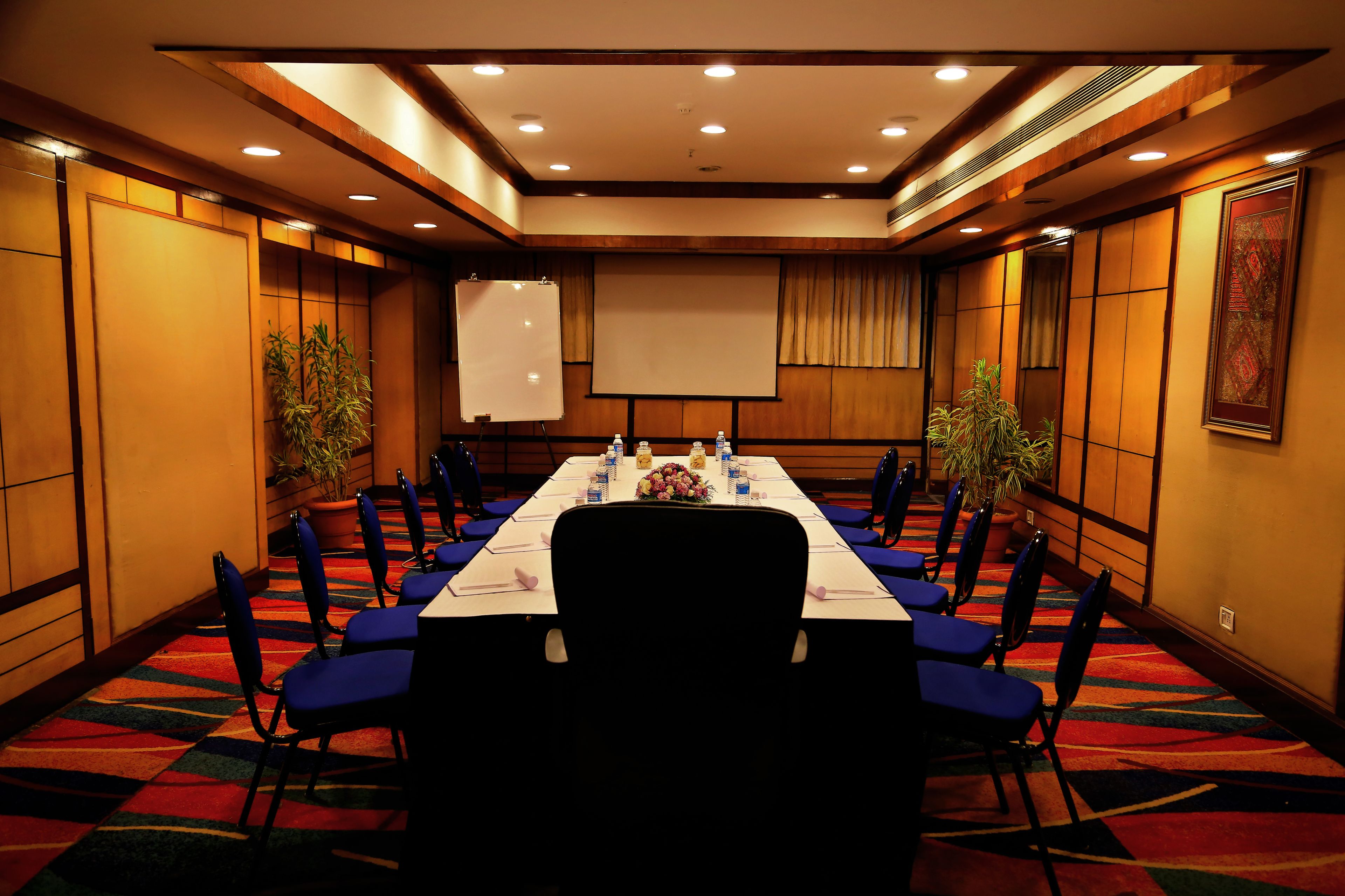 A conference room with a long table, blue chairs, a projector screen, and potted plants.