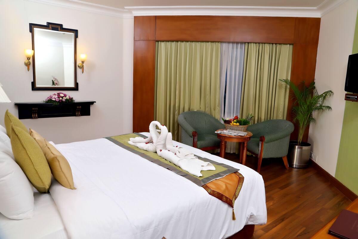 Ein Hotelzimmer mit einem ordentlich bezogenen Bett, einem grünen Polsterstuhl, einem kleinen Tisch mit einer Pflanze und einem an der Wand montierten Fernseher.