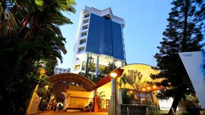 Entrada de hotel com galeria coberta, um edifício grande ao fundo e um balcão de recepção bem iluminado.