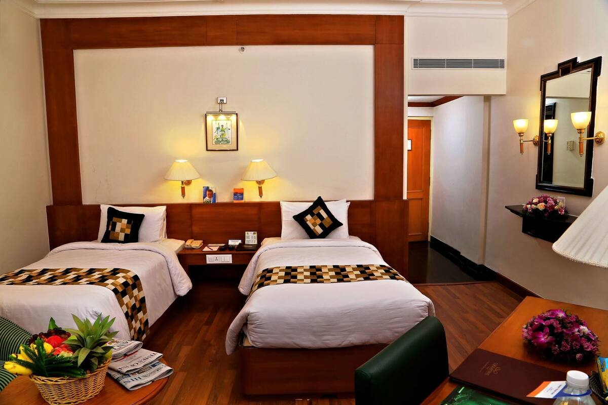 Ein Hotelzimmer mit zwei Betten, einem Schreibtisch, einem Stuhl, einem kleinen Tisch mit Blumenvase und einem Spiegel an der Wand.