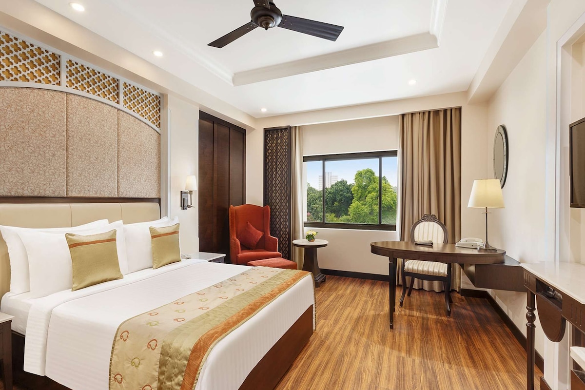 A hotel room with a bed, a desk, a chair, a ceiling fan, and a window with a view of trees.