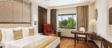 A hotel room with a bed, a desk, a chair, a ceiling fan, and a window with a view of trees.