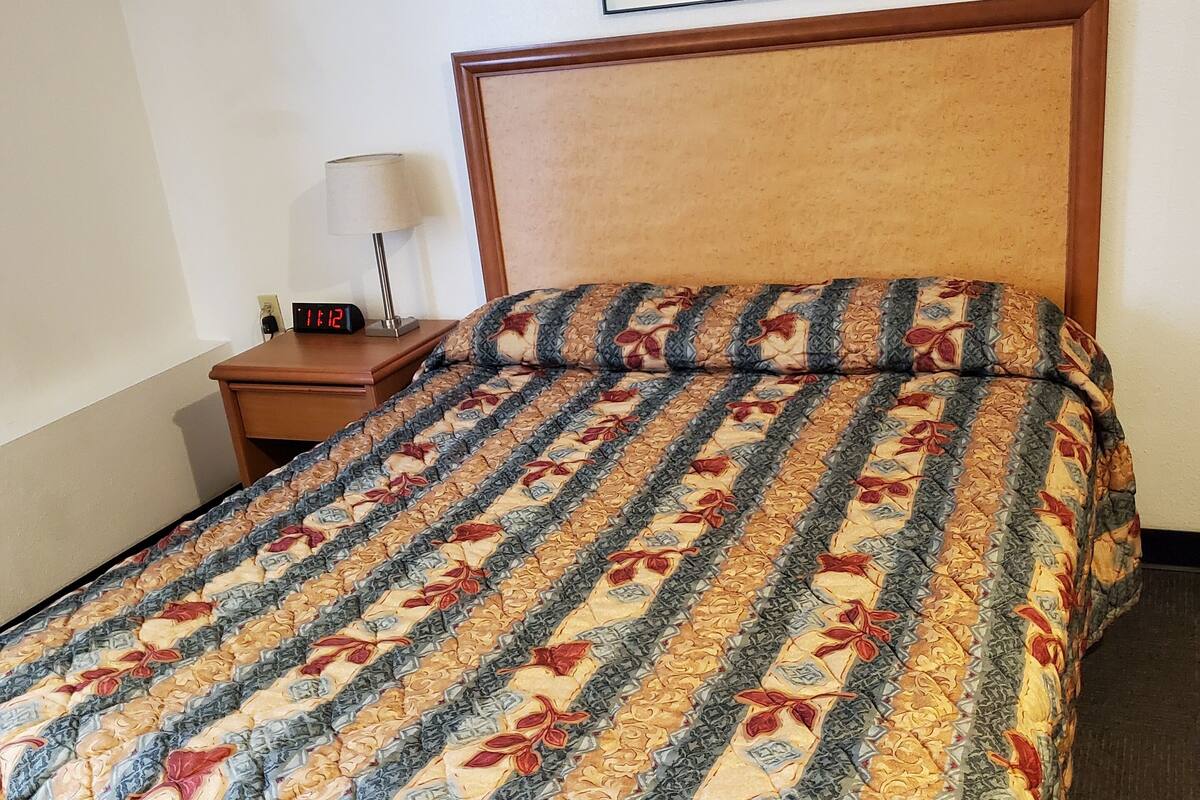 A neatly made bed with a patterned bedspread, a wooden headboard, a nightstand with a lamp, and a framed picture on the wall.
