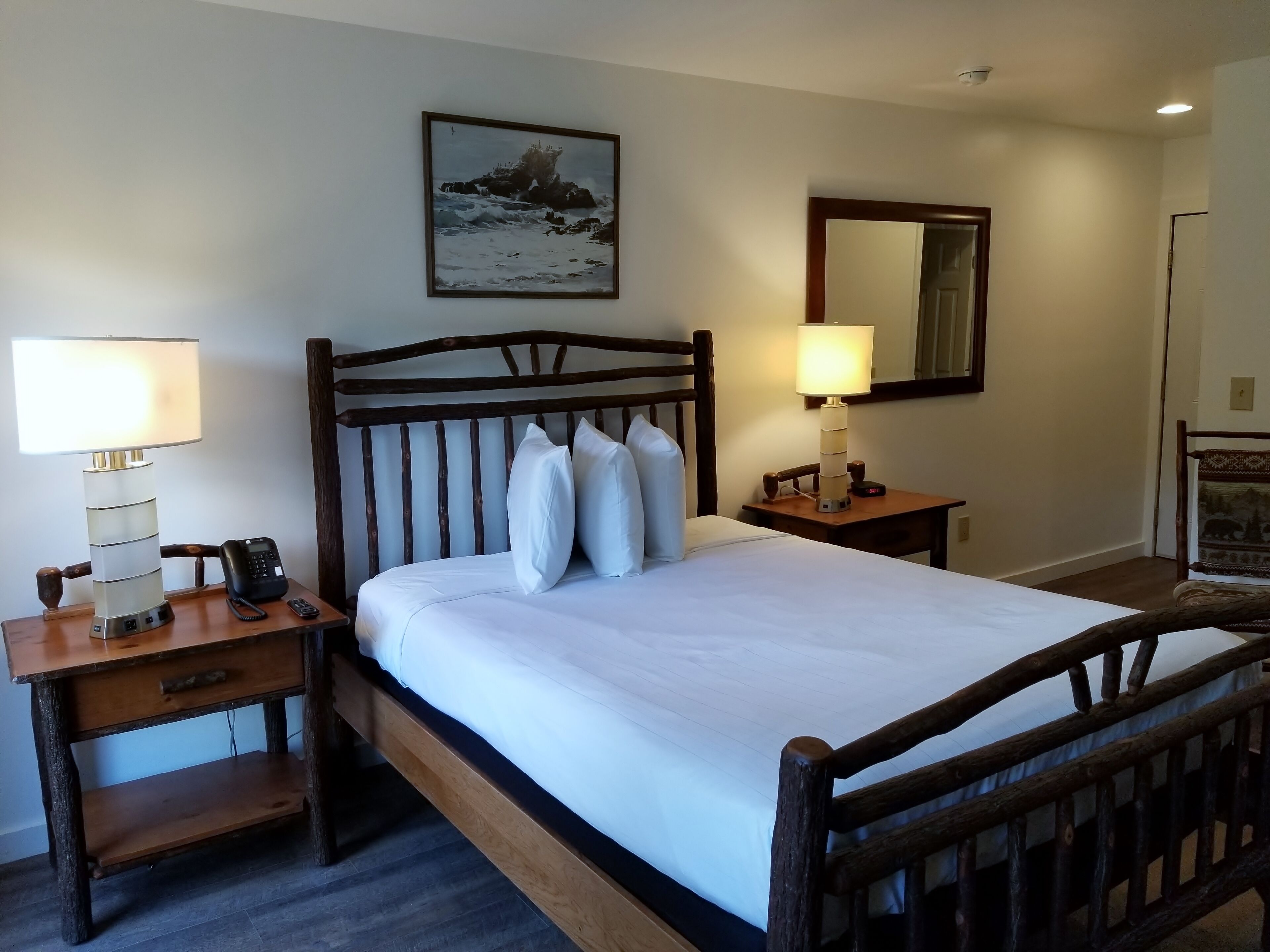 A neatly made bed with a wooden headboard, a bedside table with a lamp, a phone, and a small drawer. There is a framed picture on the wall above the bed.