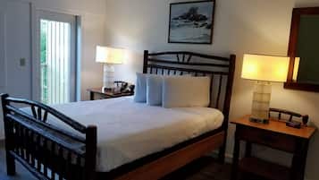 Una cama bien hecha con ropa de cama blanca, un cabecero de madera, dos mesitas de noche con lámparas, un cuadro enmarcado en la pared y una puerta que da a un balcón.