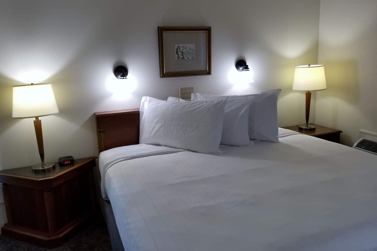 A hotel room with a neatly made bed, two bedside tables with lamps, a framed picture on the wall, and a digital clock on one of the tables.