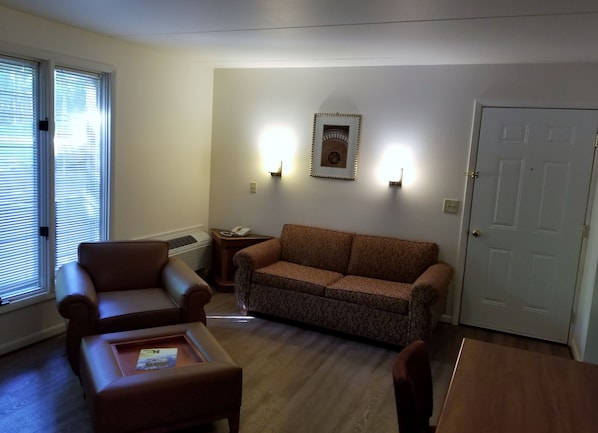 Una sala de estar con un sofá marrón, un sillón marrón y una mesa de centro de madera. Hay un teléfono montado en la pared y un cuadro enmarcado sobre el sofá.