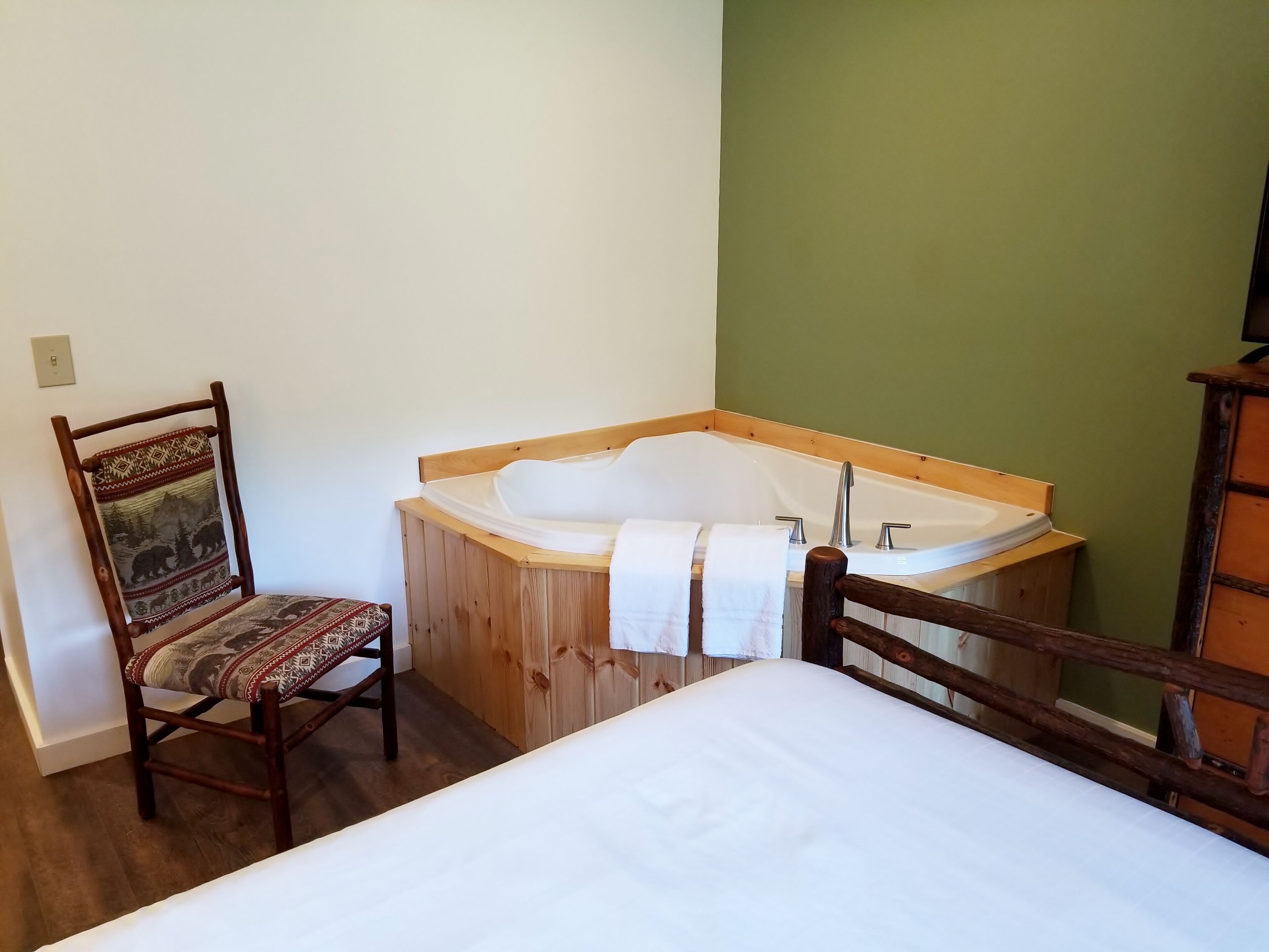 A room with a wooden bed, a wooden chair with a patterned cushion, and a large bathtub with a wooden frame.