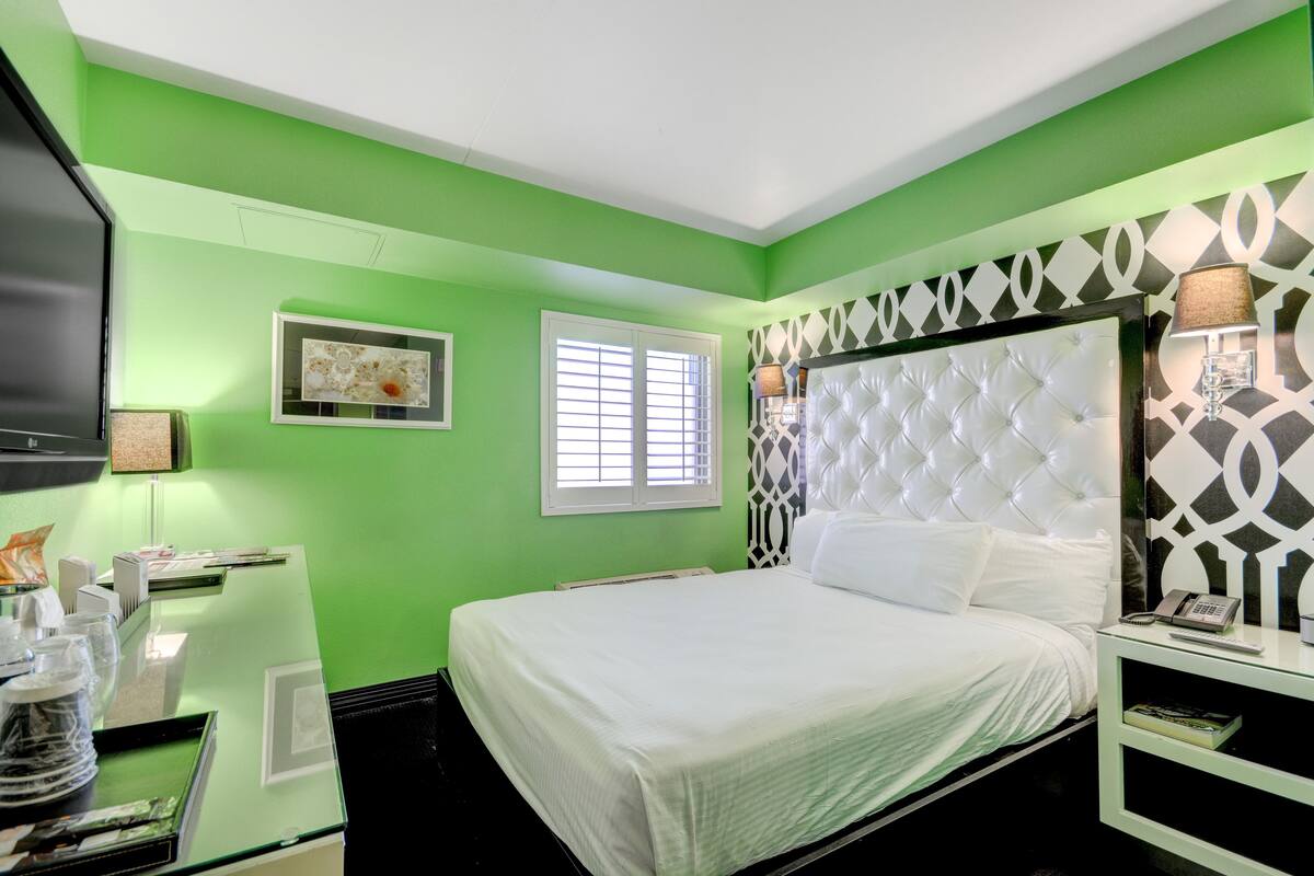 A hotel room with a green wall, a white bed, a television, a window with blinds, and a decorative wall panel.