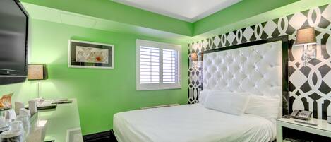 A hotel room with a green wall, a white bed, a television, a window with blinds, and a decorative wall panel.