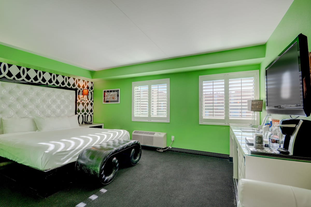A hotel room with a green wall, a large bed, a flat-screen TV, and a desk with a coffee machine.