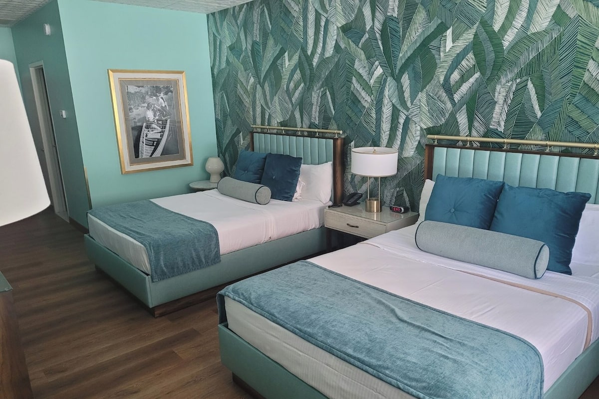 A hotel room with two beds, a framed picture on the wall, and a tropical leaf-patterned wallpaper.