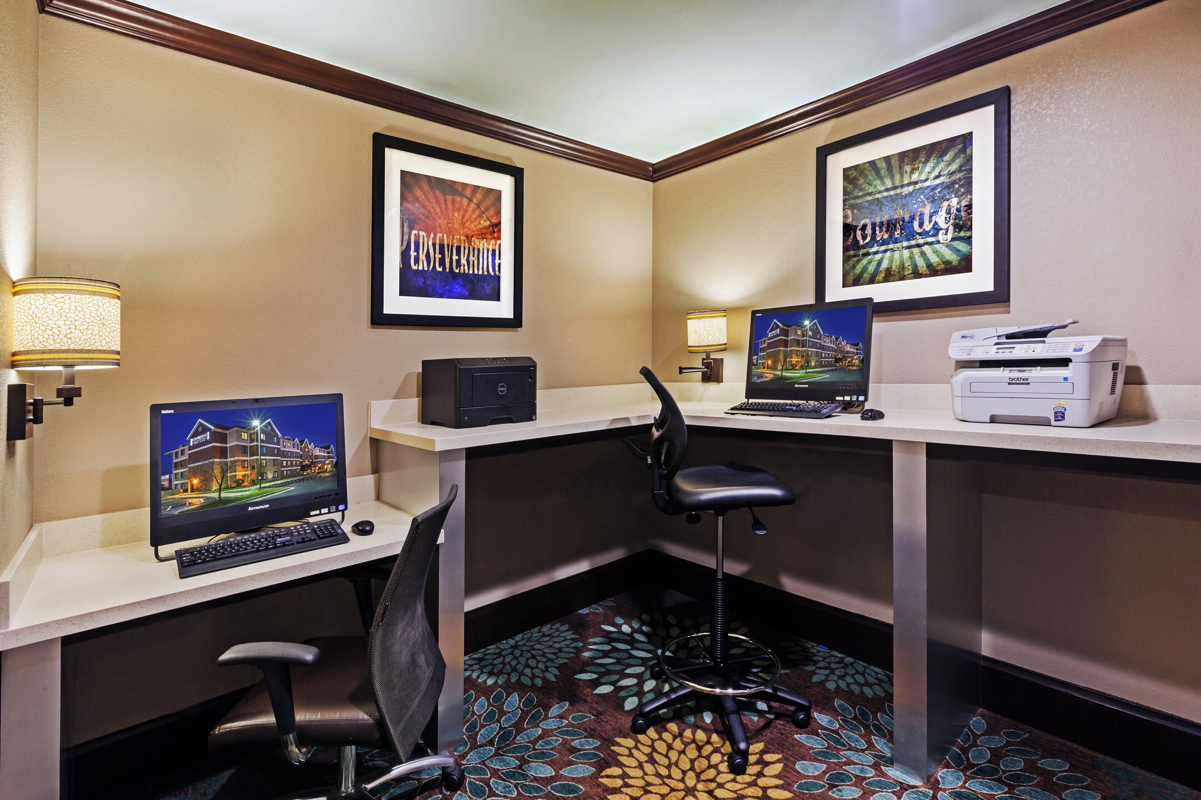 A hotel room with a desk, computer, printer, and two framed pictures on the wall.