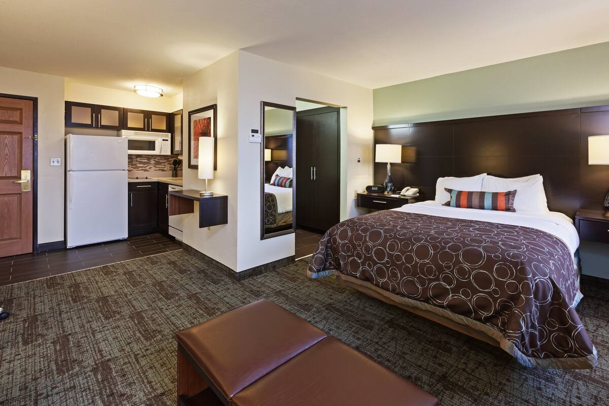 Een hotelkamer met een bed, nachtkastje, lamp, spiegel, een klein kitchenette en een badkamers deur.
