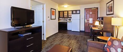 A compact hotel room with a flat-screen TV, a desk with a lamp, a kitchenette, and a sofa with a patterned cushion.