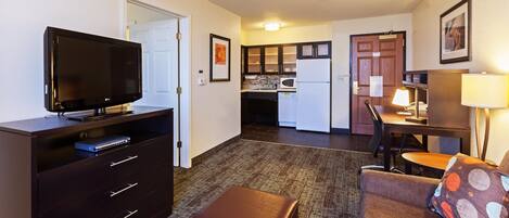 A compact hotel room with a television, a desk, a chair, a sofa, and a kitchenette.