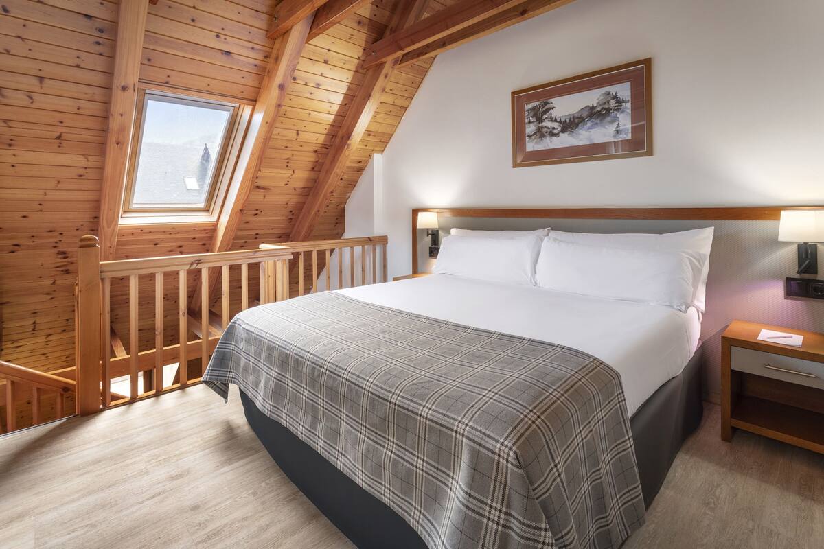 A bedroom with a wooden ceiling, a bed with white linens, a plaid blanket, a nightstand, and a framed picture on the wall.