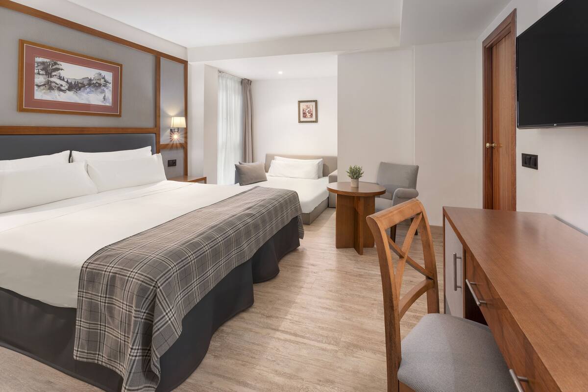 A hotel room with a large bed, a desk, and a chair. There is a television on the wall and a window with curtains.