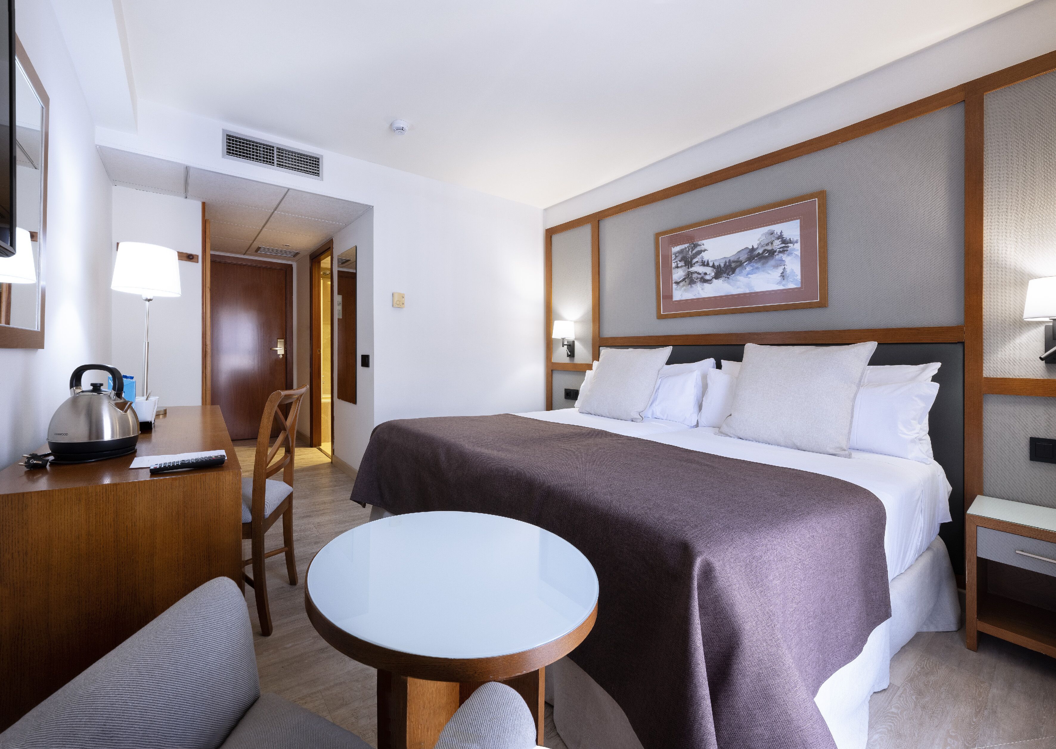 A hotel room with a bed, a desk with a kettle, a chair, a round table, and a painting on the wall.