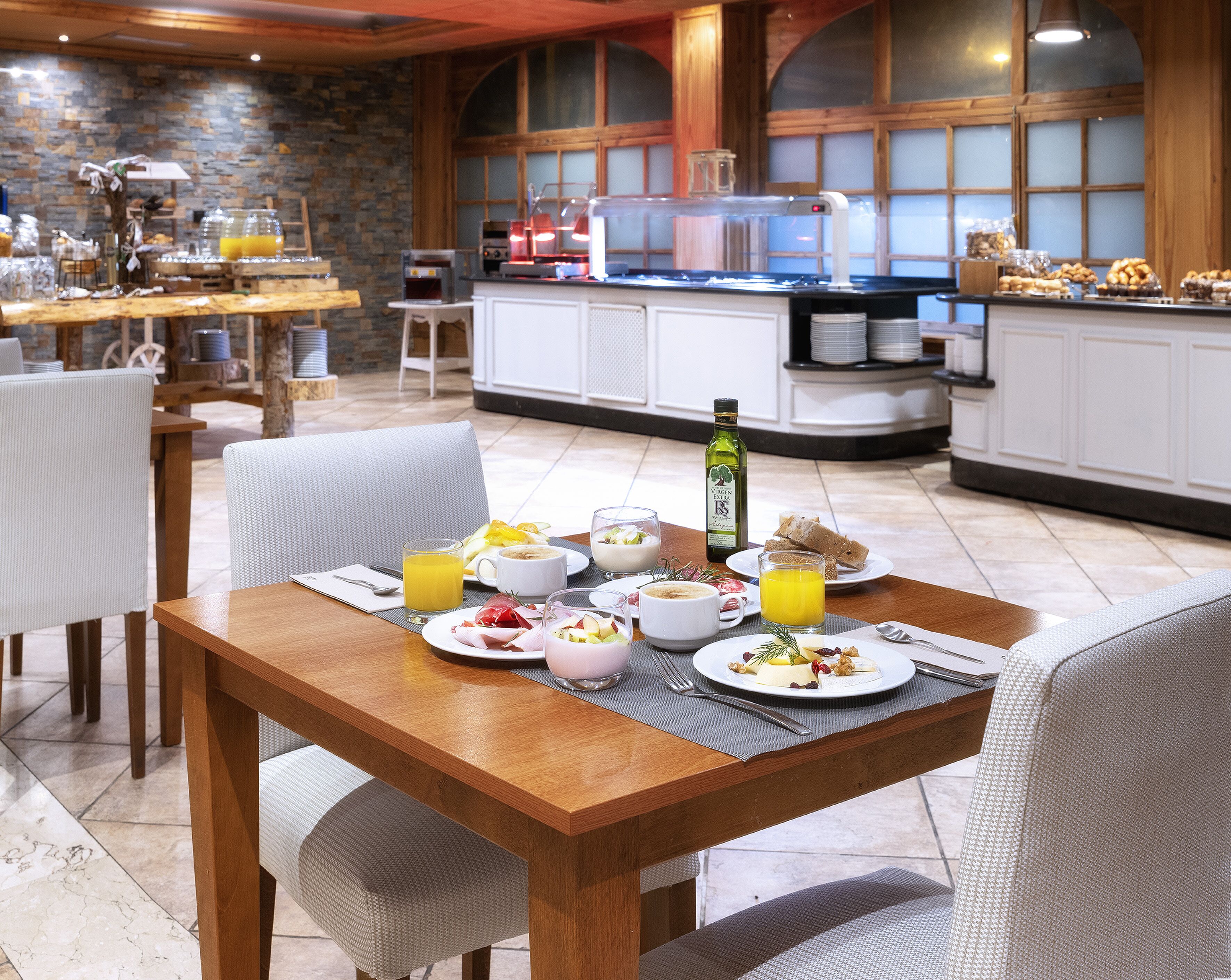 Un desayuno servido en una mesa de madera con sillas y una variedad de alimentos, como zumo, café y bollería.