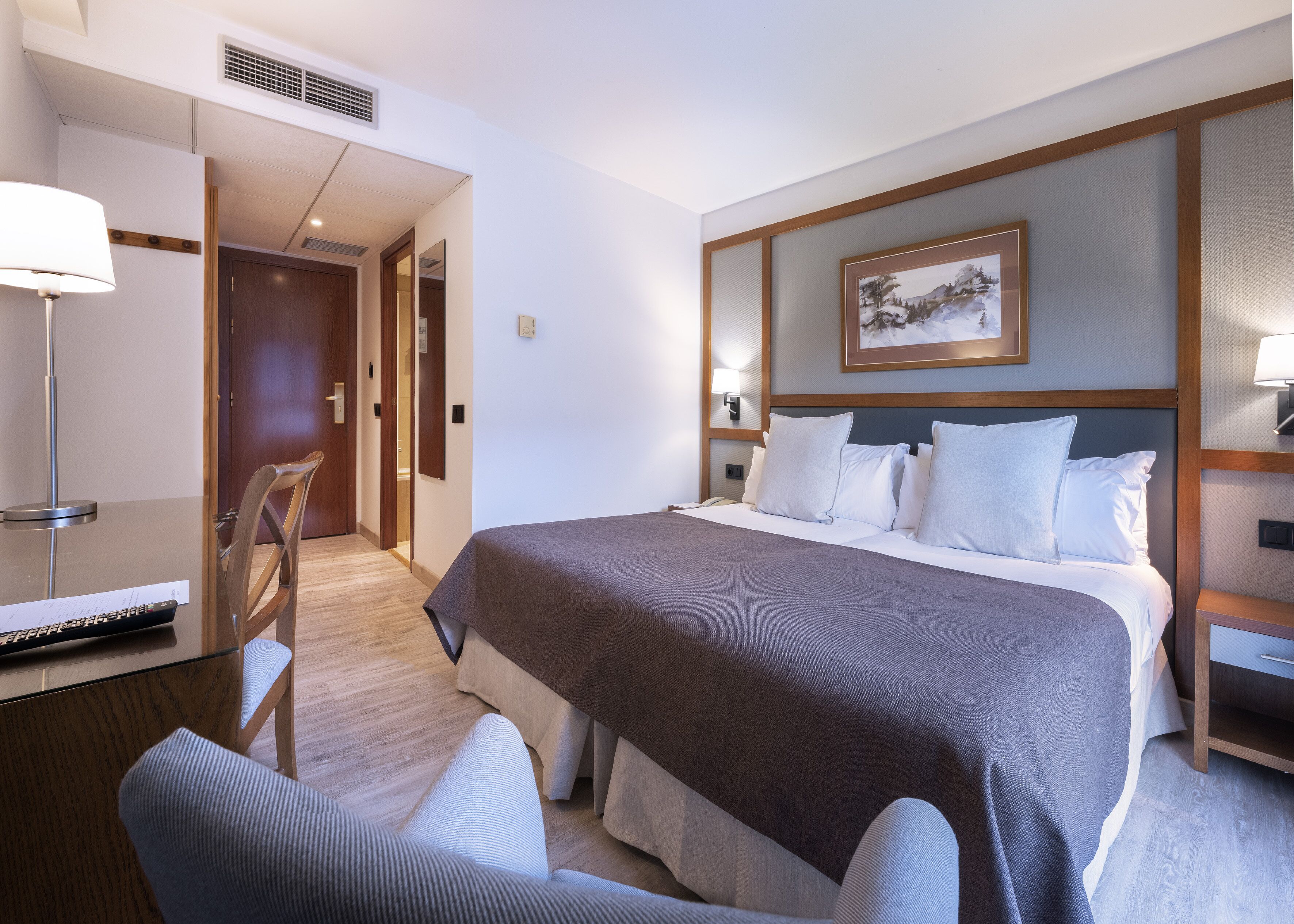Ein Hotelzimmer mit einem Bett, einem Schreibtisch mit Lampe, einem Sessel und einem Bild an der Wand.