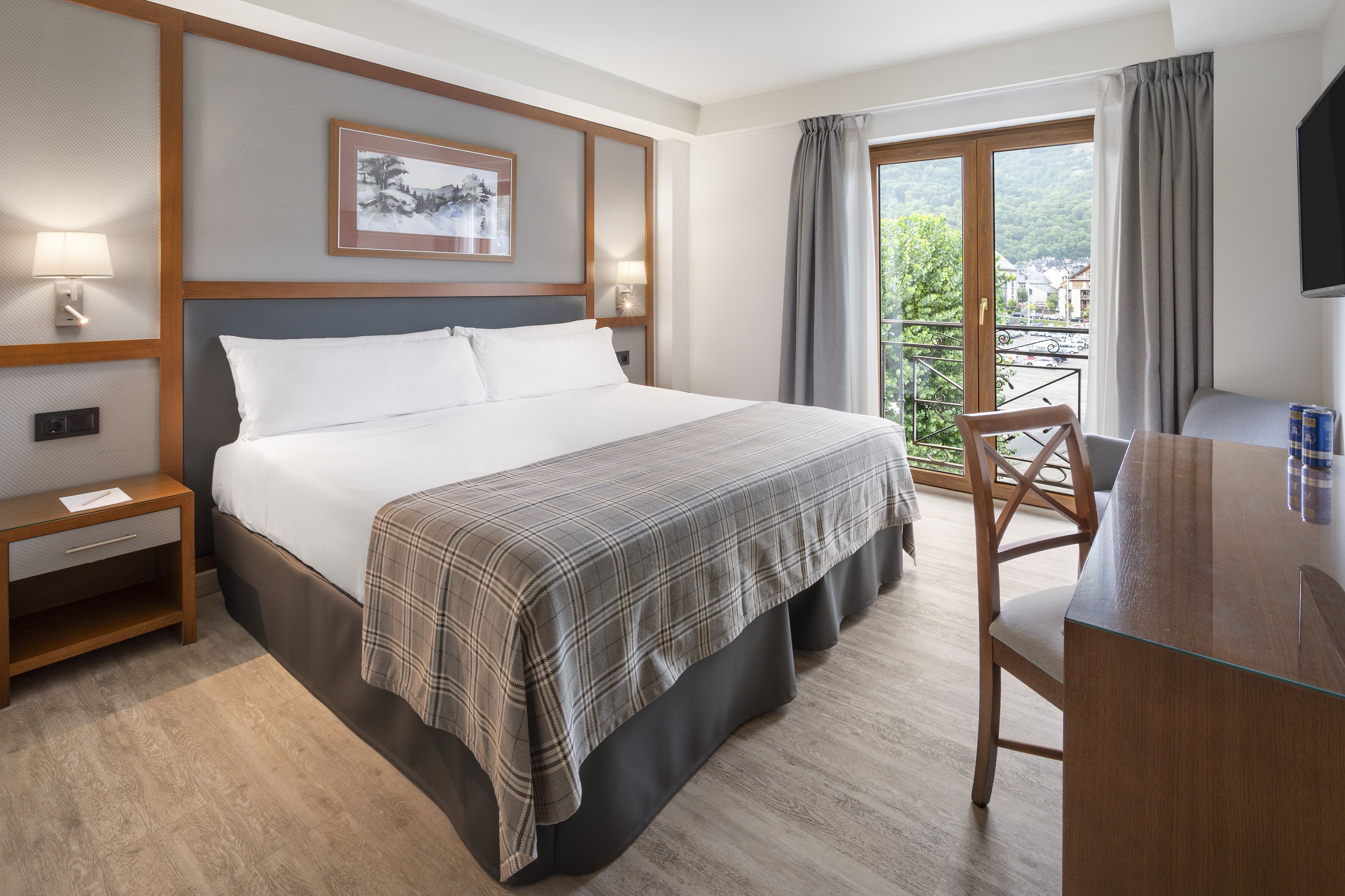 A hotel room with a large bed, a desk, a chair, a TV, and a balcony with a view of greenery.