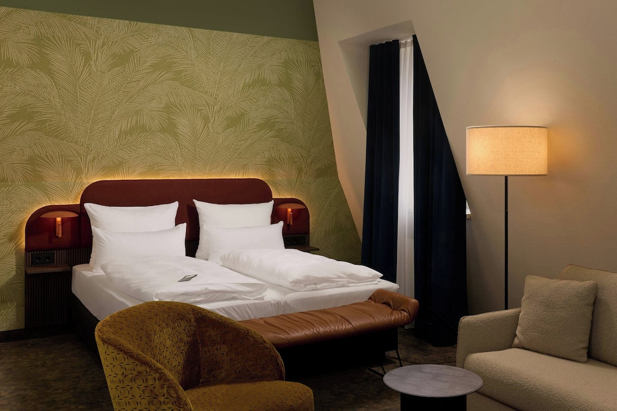 Une chambre d’hôtel avec un lit, une chaise, un canapé, une lampe et un rideau.