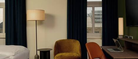 A hotel room with a bed, a desk with a laptop, a chair, a lamp, and a view of a building through the window.