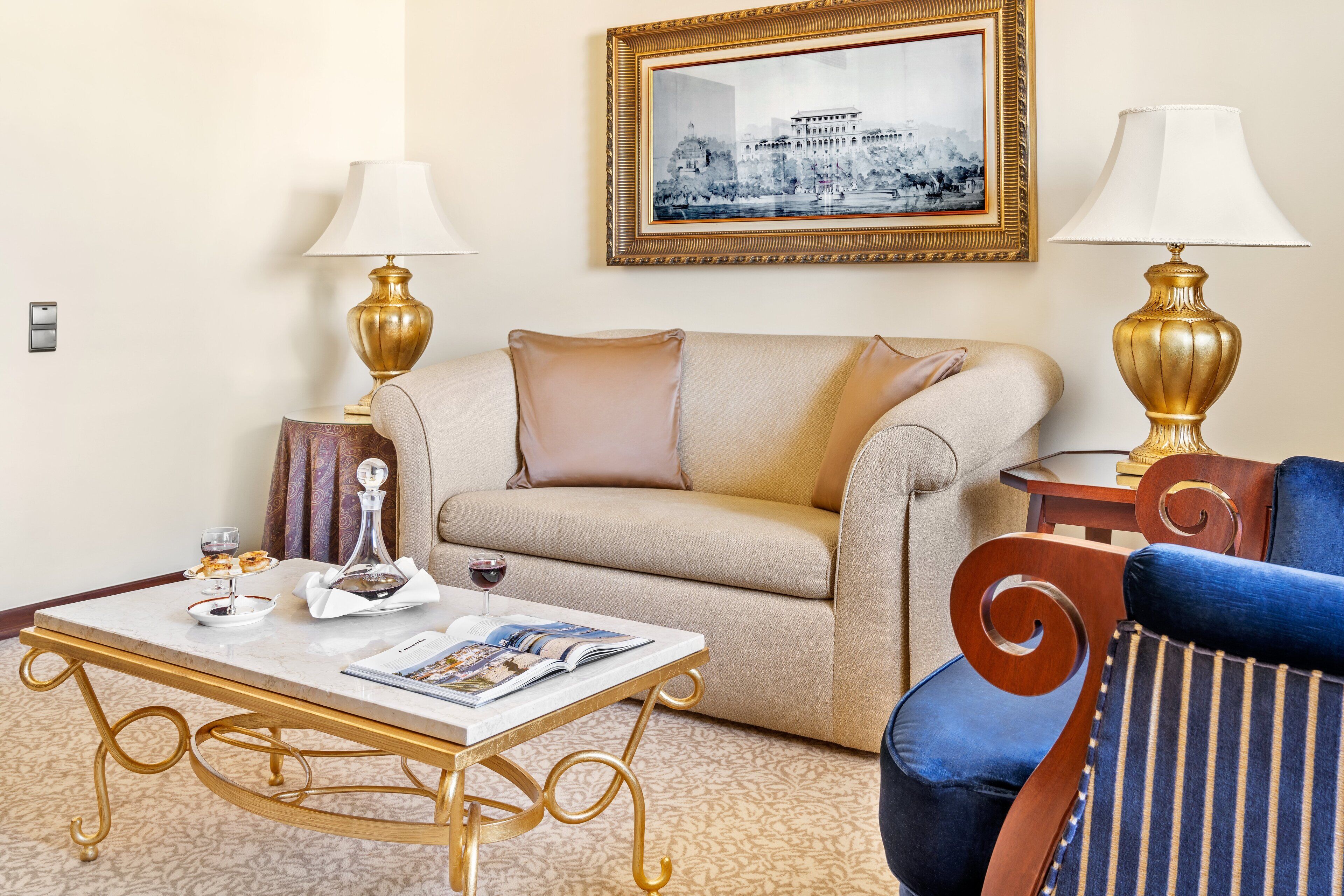 A hotel room with a beige sofa, a blue armchair, a gold-framed picture on the wall, and a small table with a magazine and a wine glass.