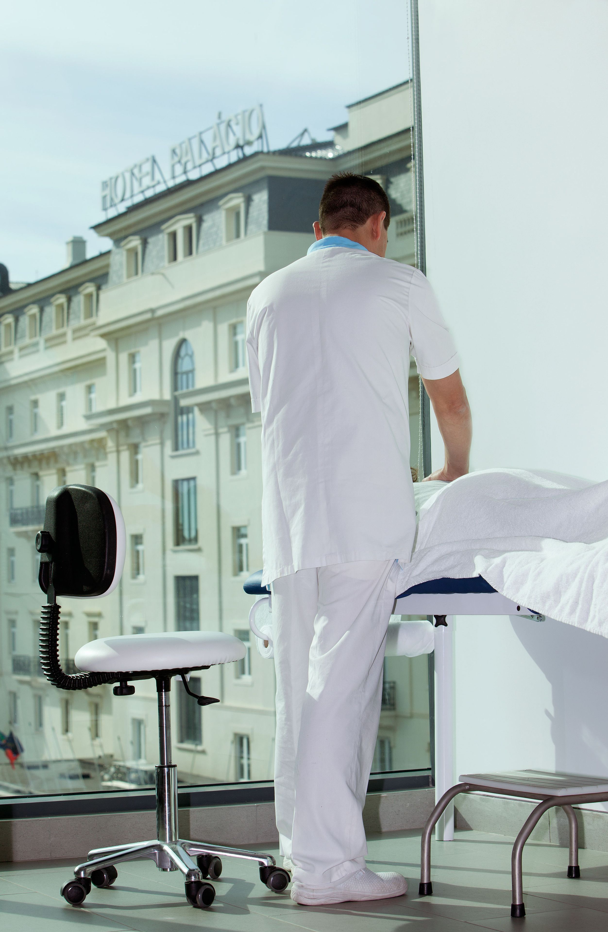 Una persona con bata blanca de hospital junto a una cama, con vista a un edificio que muestra el cartel 'HOTEL PALACE'.